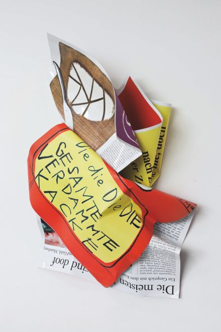 Wandskulptur von Olaf Metzel. Aluminium, das beidseitig bunt bedruckt ist und in der Form von zwei zerknüllten Blättern gefaltet ist. Auf der einen Seite ist ein Zeitungsartikel, auf der anderen steht in Handschrift: Die die Die DIE GESAMTE VERDAMMTE KACKE auf gelbem Grund, rot umrandet