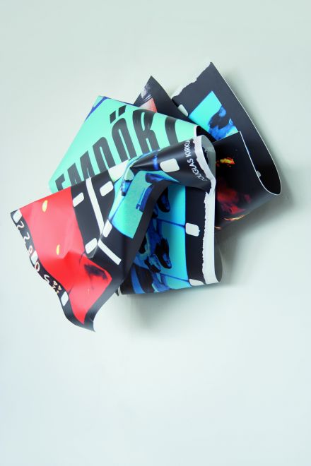 Wandskulptur von Olaf Metzel. Aluminium, das beidseitig bunt bedruckt ist und in der Form von einem zerknüllten Blatt gefaltet ist. Auf der einen Seite sind Filmstreifen, in einer Faltung ist EMPÖRT zu lesen