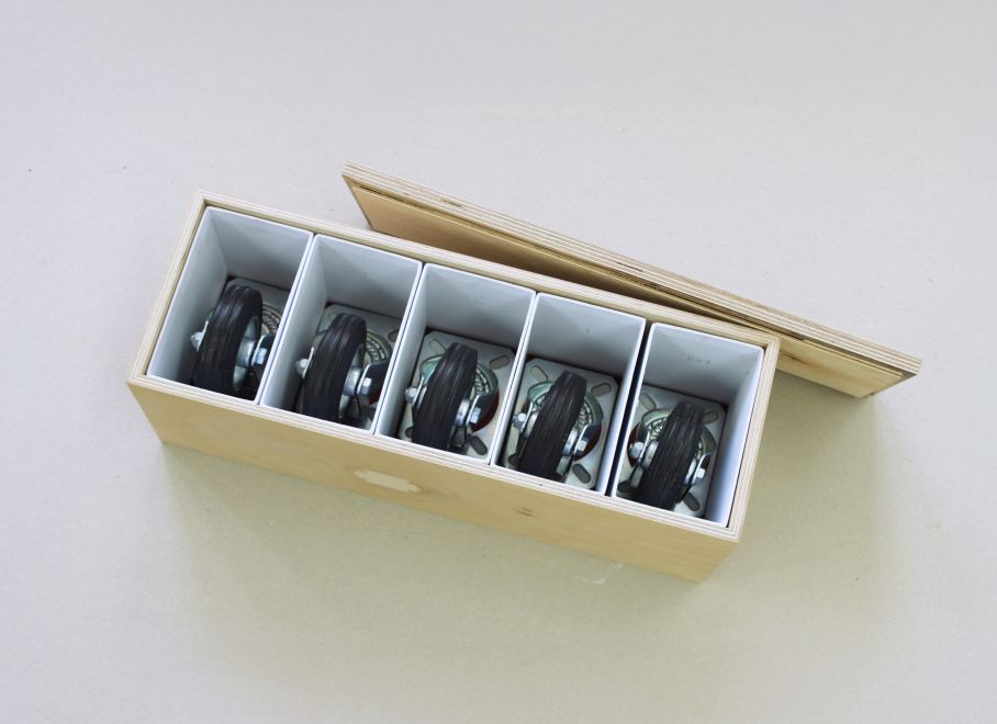 Ein länglicher Kasten aus hellem Holz ist von oben zu sehen, der Deckel steht angelehnt daneben. In dem Kasten sind 5 weiße Kartons aneinandergereiht, in denen jeweils eine Plattform Rolle liegt.