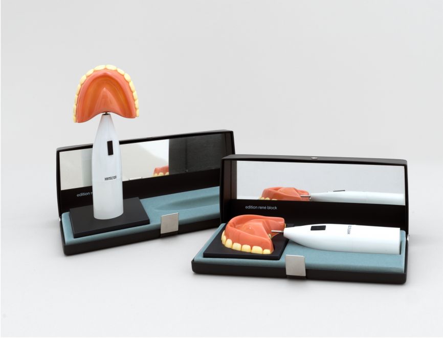 Multiple von Richard Hamilton. Die Halterung einer elektrischen Braun-Zahnbürste steht in dem zugehörigen schwarzen Etui. Anstelle der Zahnbürste ist ein Gebiss auf der Halterung. Im Etui steht: edition rene block, auf der Halterung steht HAMILTON. Daneben liegt eine zweite Edition im Etui.