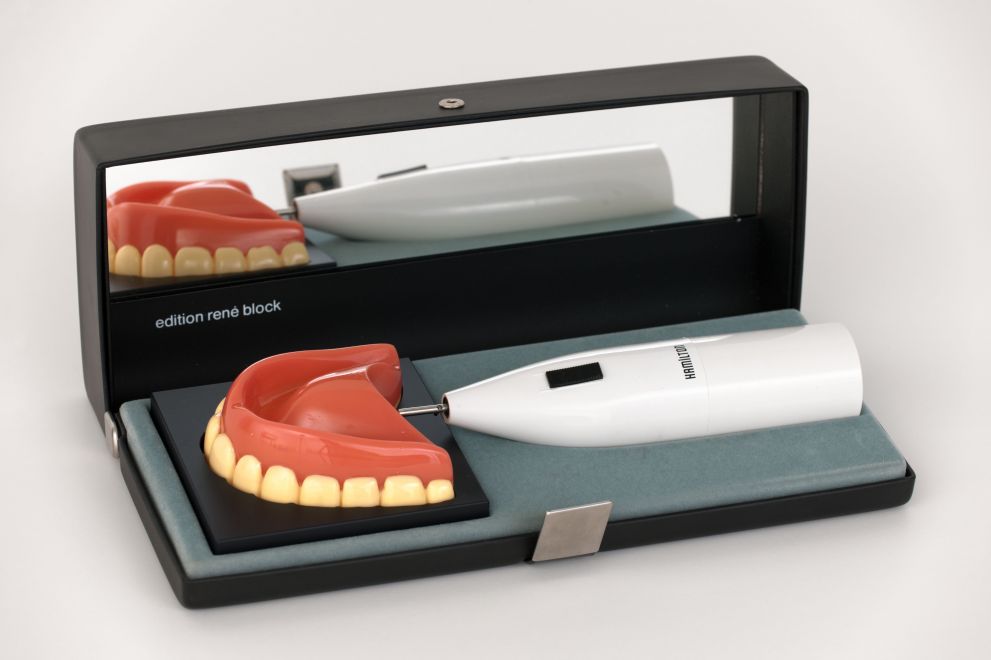 Multiple von Richard Hamilton. Die Halterung einer elektrischen Braun-Zahnbürste liegt in dem zugehörigen schwarzen Etui. Anstelle der Zahnbürste ist ein Gebiss auf der Halterung. Im Etui steht: edition rene block, auf der Halterung steht HAMILTON.