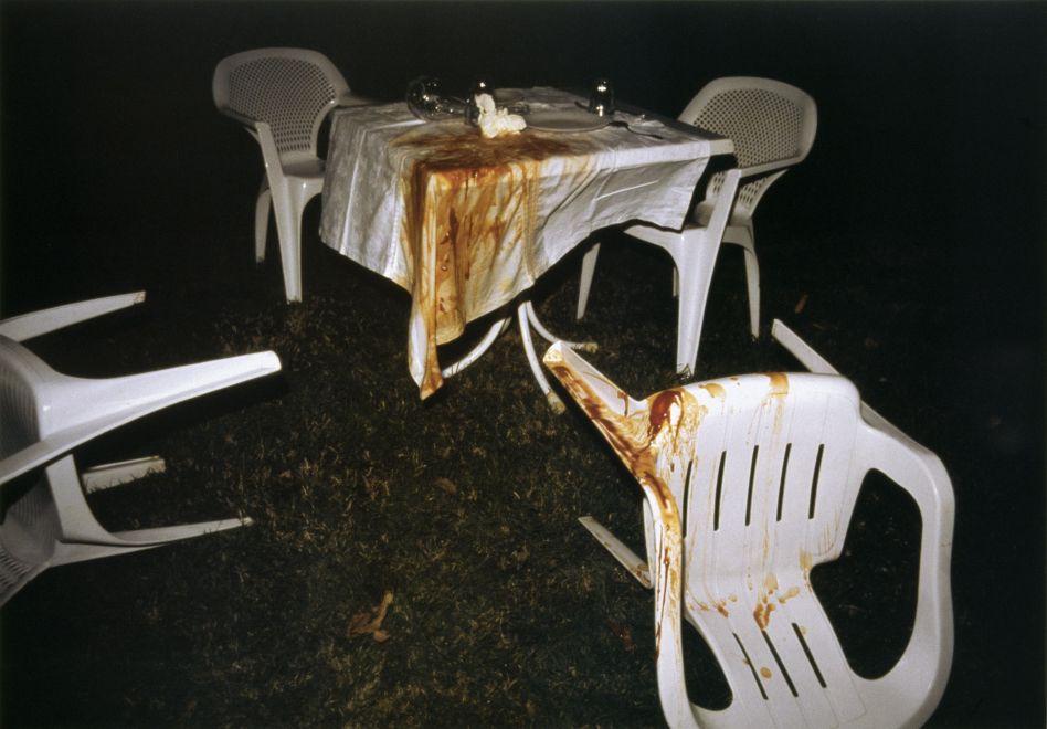 Offsetdruck von Bülent Sangar. Nachtaufnahme mit Blitz. Ein Tisch auf einer Wiese, 4 Plastikstühle, zwei davon umgeworfen. Die Tischdecke und ein Stuhl sind mit roter Sauce verschmiert. Sieht wie der Ort eines Verbrechens aus.