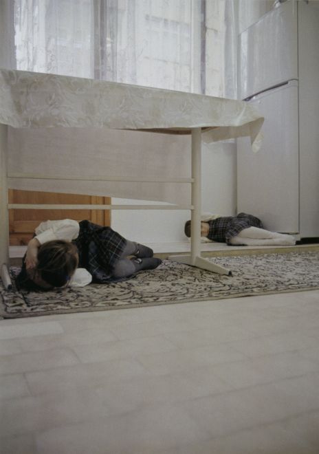 Offsetdruck von Bülent Sangar. Zwei Mädchen in Schuluniformen liegen zusammengerollt in Schutzhaltung auf dem Boden einer Küche.