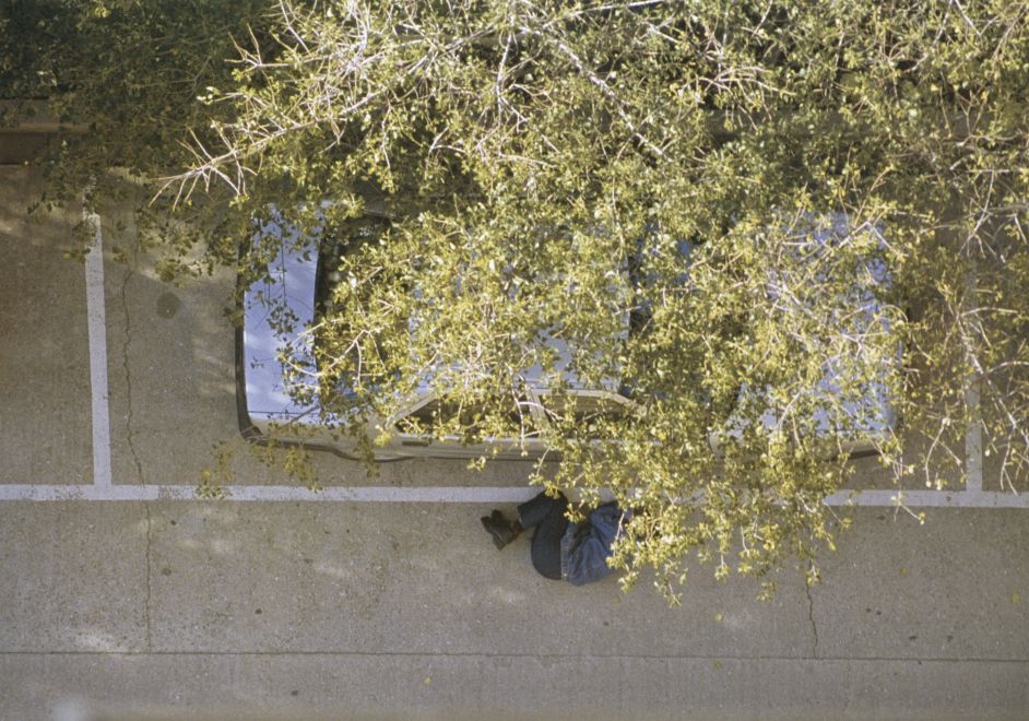 Offsetdruck von Bülent Sangar. Aufnahme aus Vogelperspektive: Blick auf einen Baum, durch die Blätter ist ein Auto zu sehen und eine Person, die zusammengerollt in Schutzhaltung auf dem Boden liegt.
