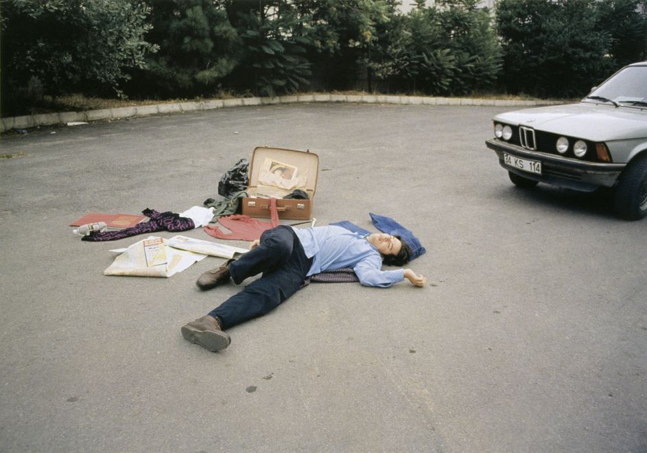 Offsetdruck von Bülent Sangar. Ein Mann liegt bewusstlos oder tot auf der Straße, neben ihr ein geöffneter Koffer, dessen Inhalt auf der Straße verstreut liegt. Am rechten Rand ein Auto