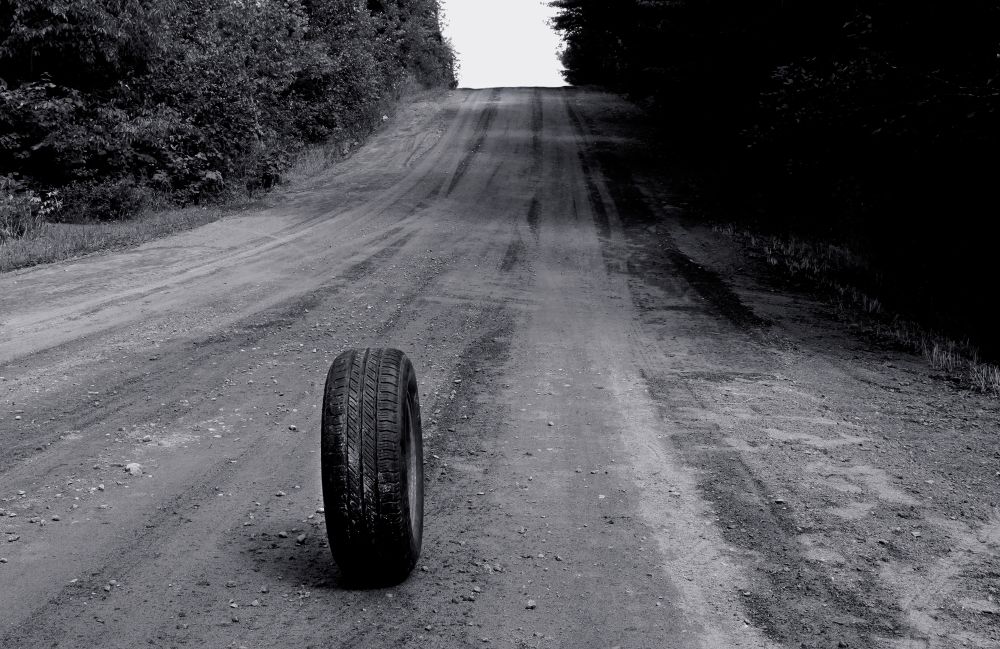 Schwarz/Weiß Fotografie von Mariana Vassileva zeigt eine breite Schotterstraße, von beiden Seiten bewachsen, die einen leichten Hügel hinaufführt. Im Vordergrund steht ein verlassener Autoreifen. Die Szene wirkt verlassen und traumartig