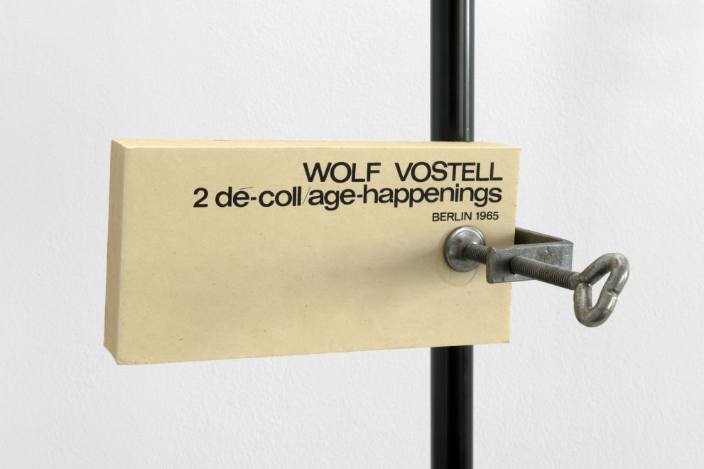 Multiple von Wolf Vostell. Längliche Karten aus vergilbten Karton sind mit einer Schraubklemme an eine Stange geklemmt. Auf der ersten Karte steht: WOLF VOSTELL. 2 de-coll/age-happenings. BERLIN 1965