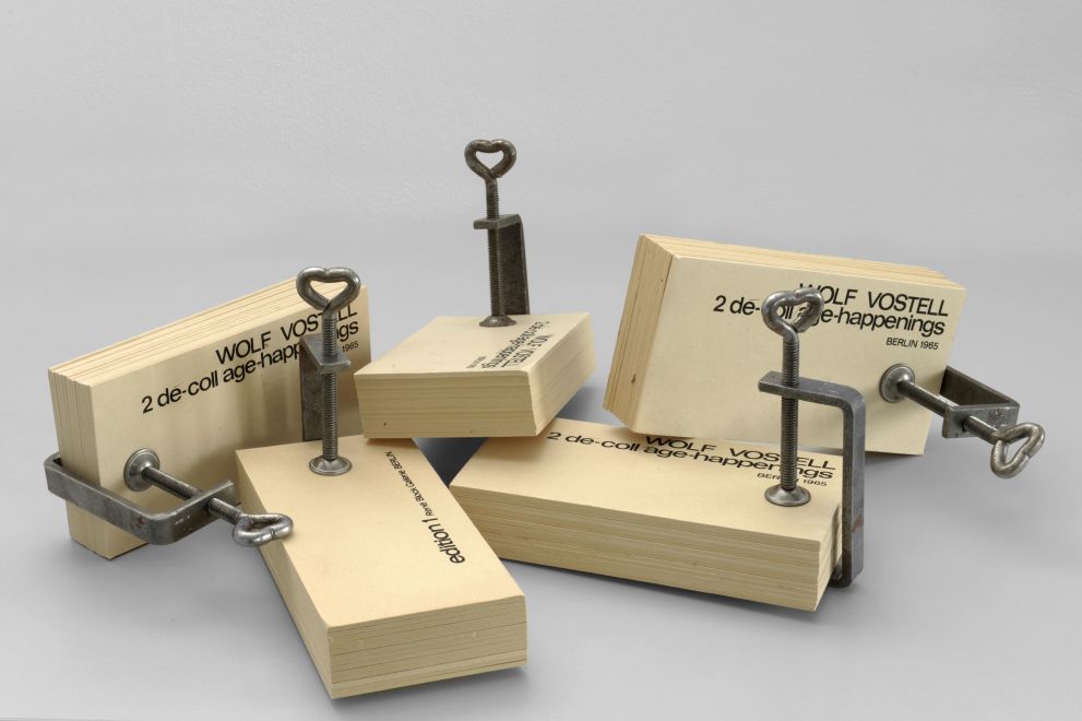 Multiple von Wolf Vostell. Längliche Seiten aus vergilbten Karton werden mit Schraubklemmen zusammengehalten. Auf der ersten Karte steht: WOLF VOSTELL. 2 de-coll/age-happenings. BERLIN 1965 Mehrere dieser Klammerbücher liegen auf einem Haufen