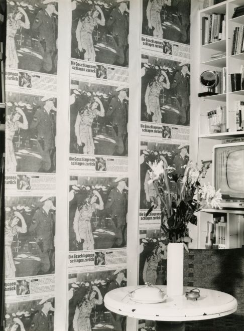 Schwarz/Weiß-Foto zeigt eine Wand, die mit der Studententapete von Vostell tapeziert ist. Als Motiv wurde eine Seite aus dem Stern über die Demonstrationen am 2.6.1967 gegen den Staatsbesuch des iranischen Schahs in Berlin unzählig wiederholt gedruckt wurden. Das Foto zeigt eine blutüberströmte junge Frau, die neben einem Polizisten läuft, im Hintergrund mehr Polizei. Vor der Wand steht ein Tisch mit einer Vase mit Blumen und ein angeschnittenes Bücherregal