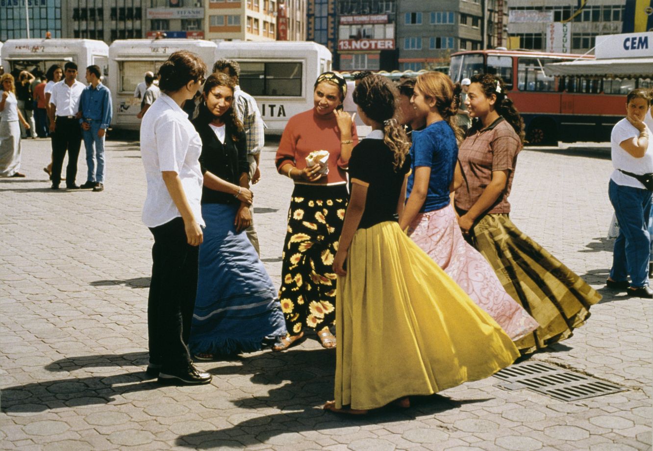 Offsetdruck von Aydan Murtezaoglu. Die Künstlerin ist formal in einem weißem Hemd gekleidet und steht in einem Kreis von Roma Frauen mit bunter Kleidung. Die Szene spielt sich auf einem belebten Kadıköy Port Square in Istanbul ab.