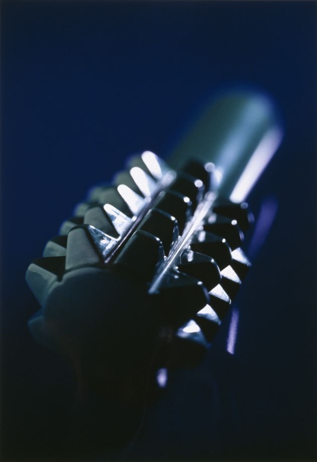 Druck der Künstlerin Ayse Erkmen. Nahaufnahme eines Landminen-Modells: ein runder, länglicher Gegenstand mit einer Art Griff und dumpfen Zacken vor dunkelblauem Hintergrund. Starke Tiefenunschärfe und Schlaglicht.