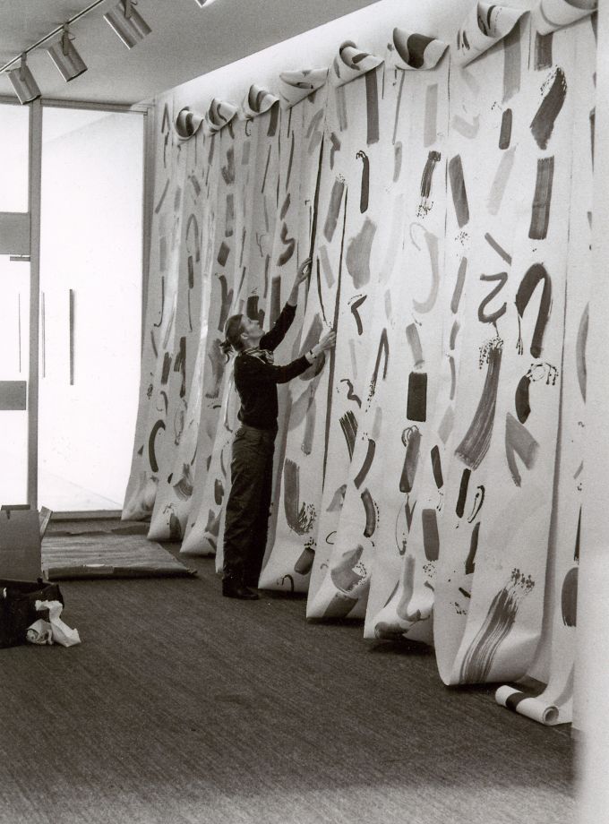 b/w photo shows Eva-Maria Schön installing her work. A long canvas strip with brushstrokes is installed in many loops over a wall