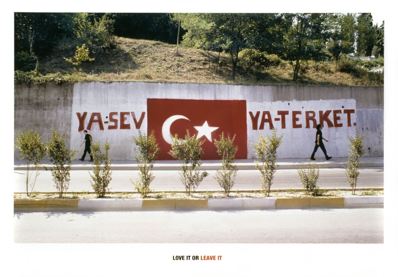 Eine Grafik vom Künstler Halil Altindere. Ein Grafitto an einer Straßenmauer zeigt die türkische Flagge und die Worte YA SEV YA TERKET, englisch Love it or leave it, in großen Buchstaben. Zwei Männer gehen auf dem Bürgersteig in entgegengesetzte Richtungen.