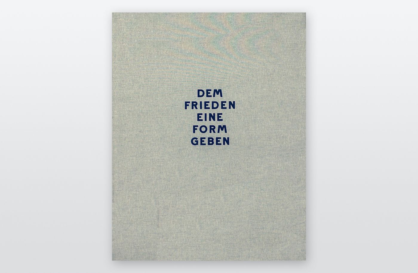 Eine graue Leinenkassette liegt auf grauem Hintergrund. Auf der Kassette steht in schwarz: DEM FRIEDEN EINE FORM GEBEN
