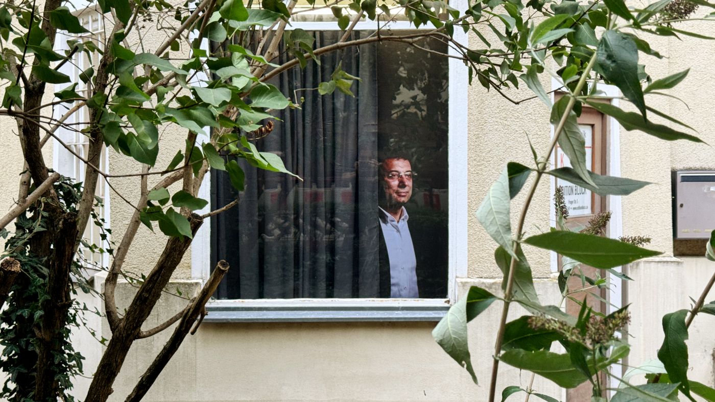 A view of a window with a gray curtain, behind which a life-size photo of Ekrem Imamoglu peeks out.
