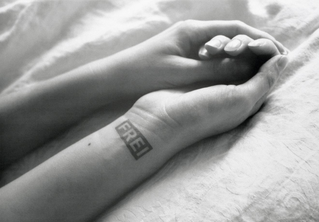 Offsetdruck von Sejla Kameric, Ein schwarz-weiß Foto von den Unterarmen und Händen einer Frau auf einem Leinentuch. Die Hände sind ineinander gelegt. Auf der Innenseite des rechten Handgelenks ist ein Stempel: FREI